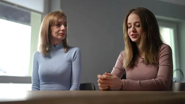 Svitlana, una mujer de cabello castaño claro que viste un suéter azul pálido de punto canalé, mira a su hija, quien mira hacia abajo, a sus manos, mientras están sentadas juntas en una entrevista.