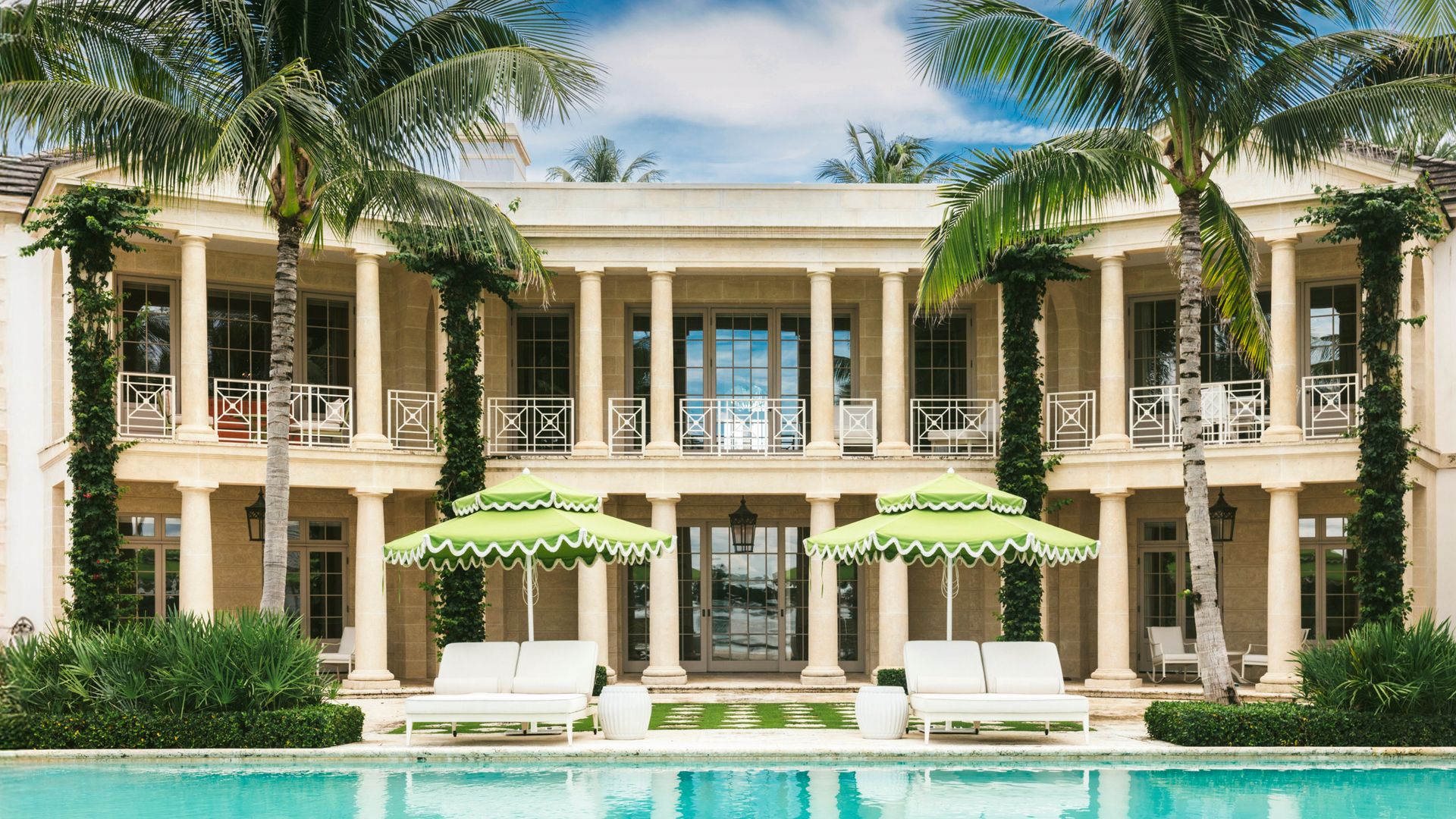 Fachada de una mansión con "infinity" pool y tumbones blancas con sombrillas verdes y altas palmeras 