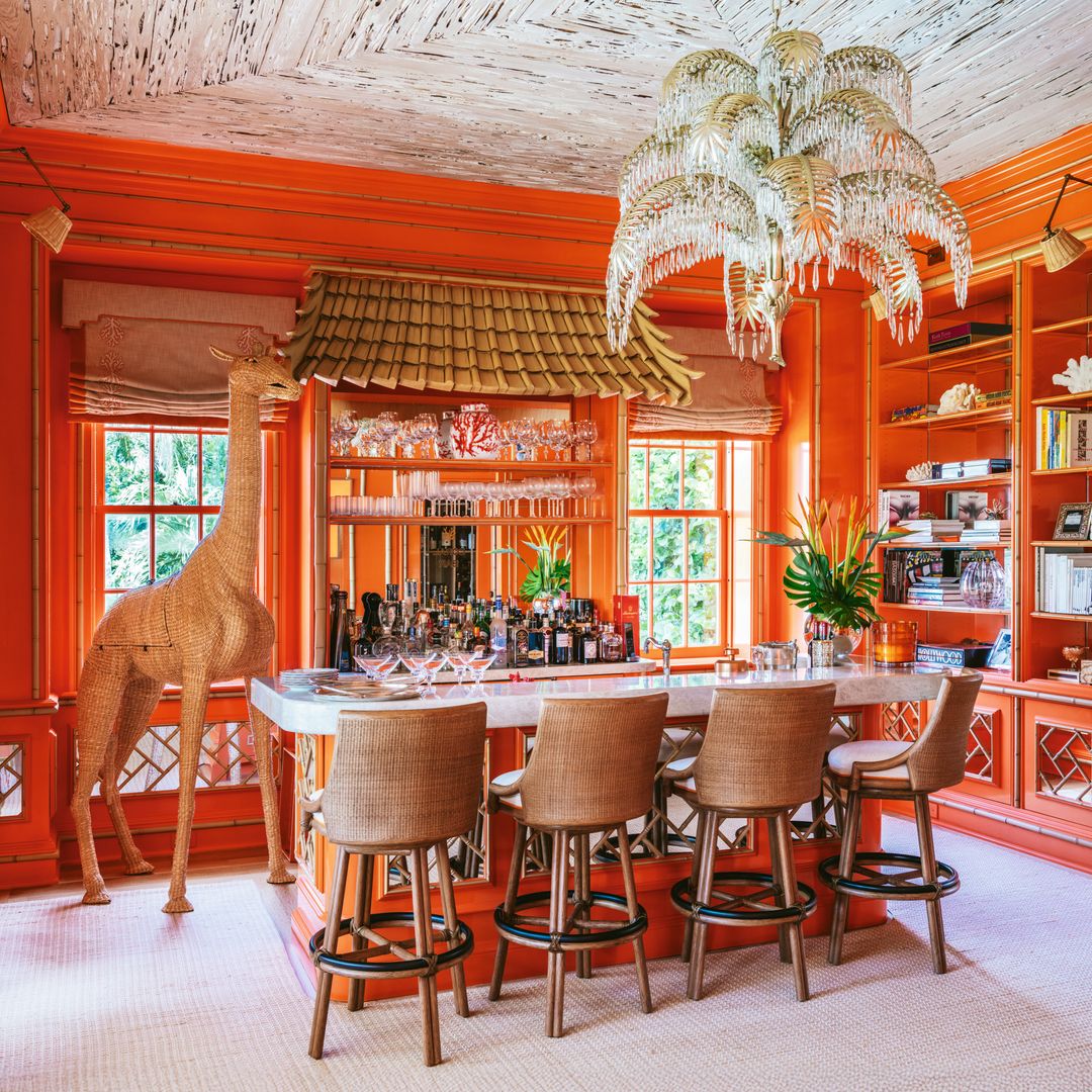 Sala de bar naranja con barra, sillas, lámpara de plumero, jirafa decorativa botellas y estanterías llenas de libros