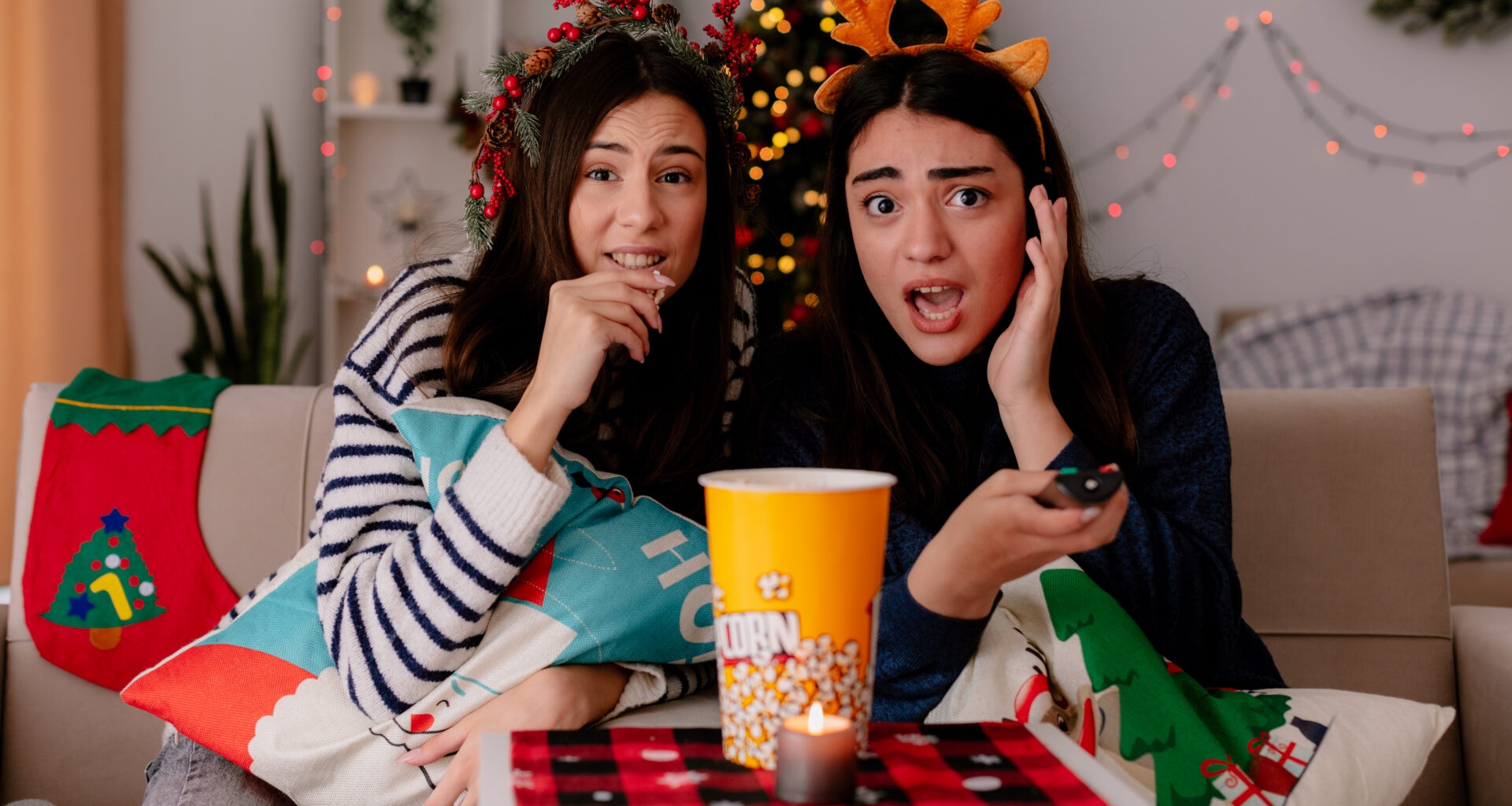 shocked pretty young girl with reindeer headband holds tv remote and looks at camera sitting on armchair with her friend eating popcorn enjoying christmas time at home
