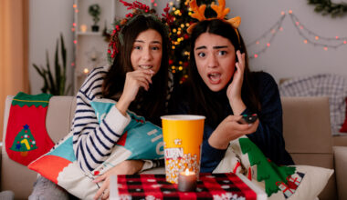 shocked pretty young girl with reindeer headband holds tv remote and looks at camera sitting on armchair with her friend eating popcorn enjoying christmas time at home