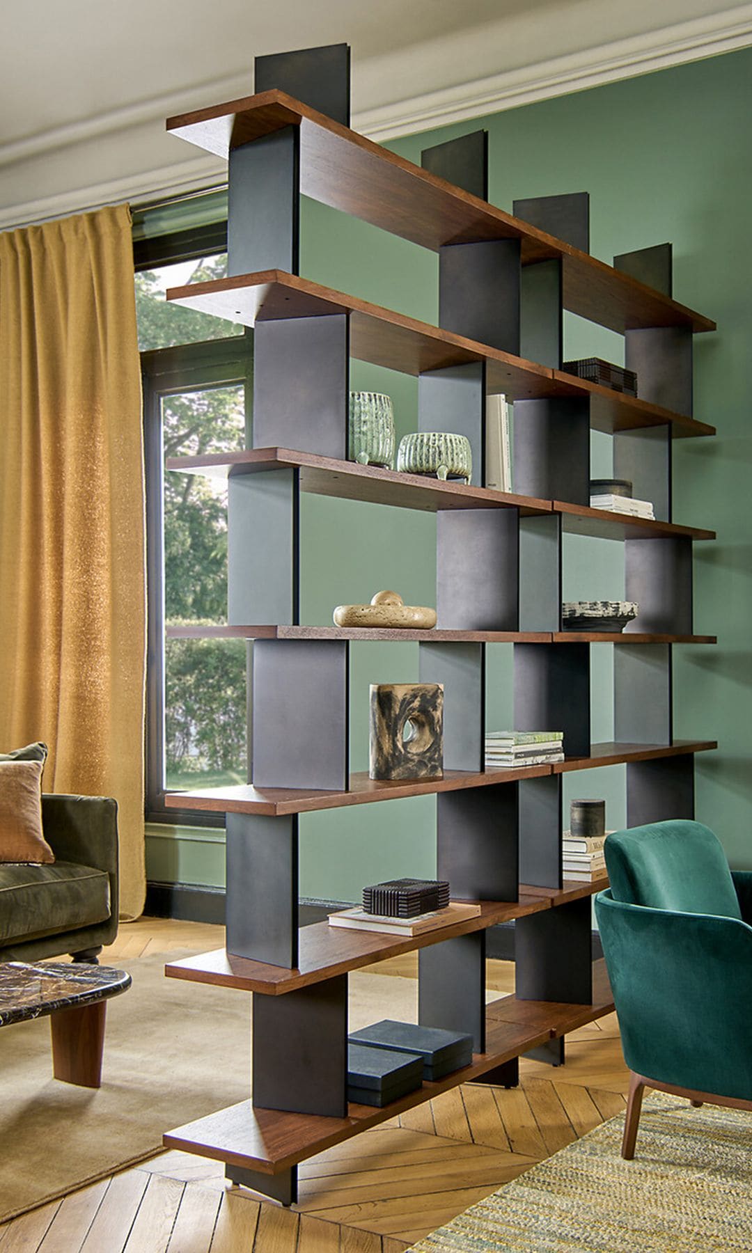Salón dividido en dos ambientes mediante una estantería abierta, sin trasera, paredes de color verde, butaca verde, sofá verde, cortinas color mostaza, alfombras