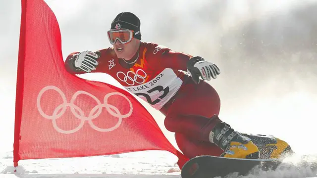 Ryan Wedding en una snowboard con el uniforme rojo de los Juegos Olímpicos de Salt Lake City, gafas de nieve, botas amarillas y un gorro negro. A su lado ondea una bandera roja con el logotipo olímpico en blanco.