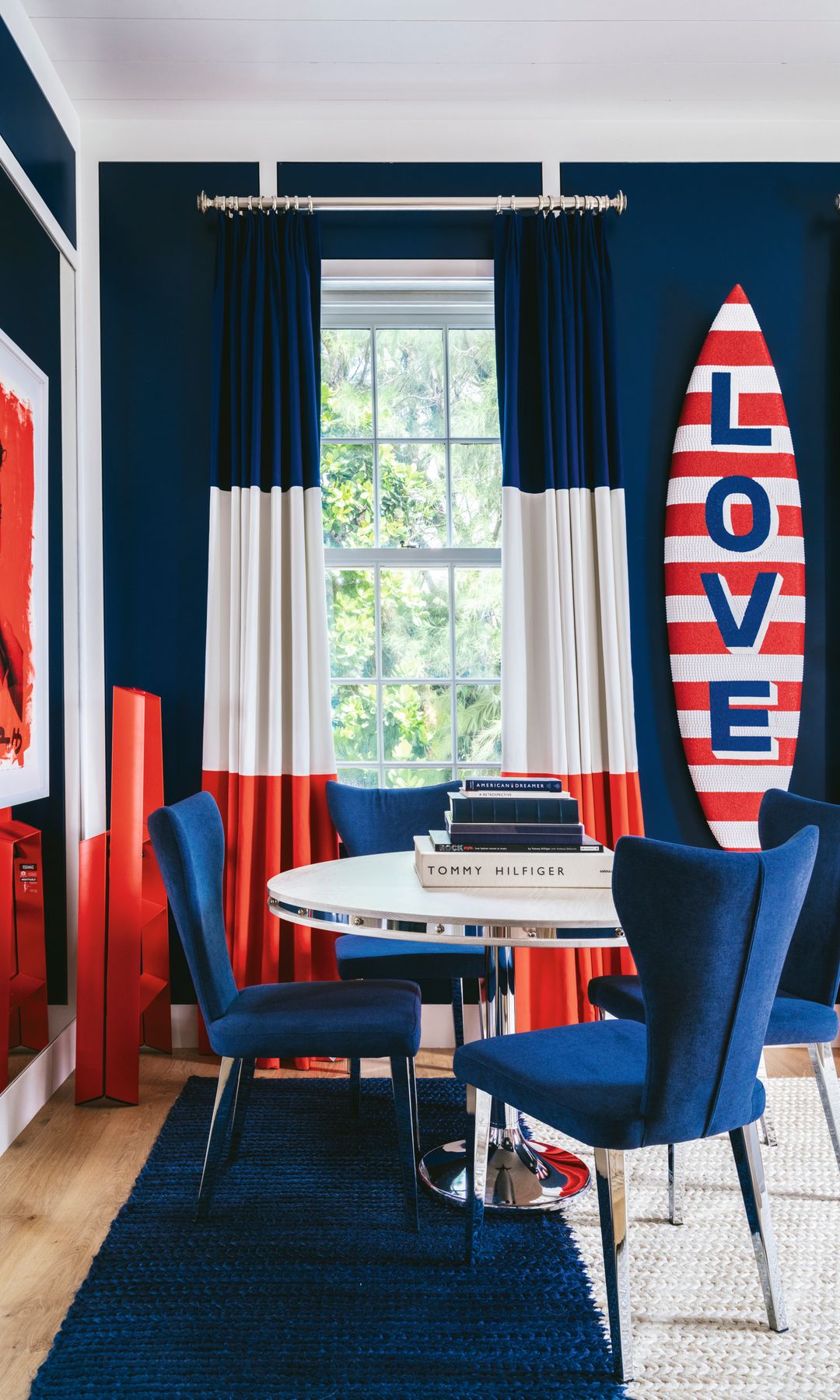 Sala con mesa redonda y sillas azules, cortinas rojas y azules, y tabla de surf de rayas rojas y blancas 