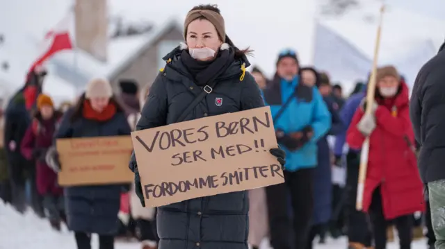 En Nuuk, Groenlandia, manifestantes abrigados con gruesos abrigos de invierno portan pancartas con la nieve de fondo. La manifestante que aparece en primer plano tiene la boca tapada con cinta adhesiva.