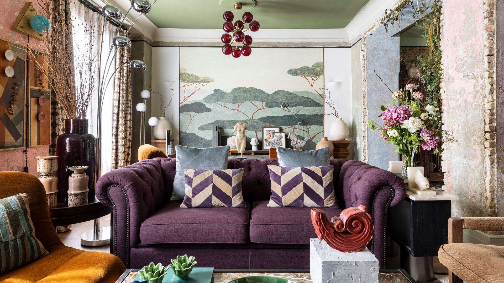 Salón maximalista con mural en las paredes, sofá morado con cojines del mismo tono en motivos geométricos, lámpara de techo con bolas rojas, jarrón con flores, falso techo de color verde