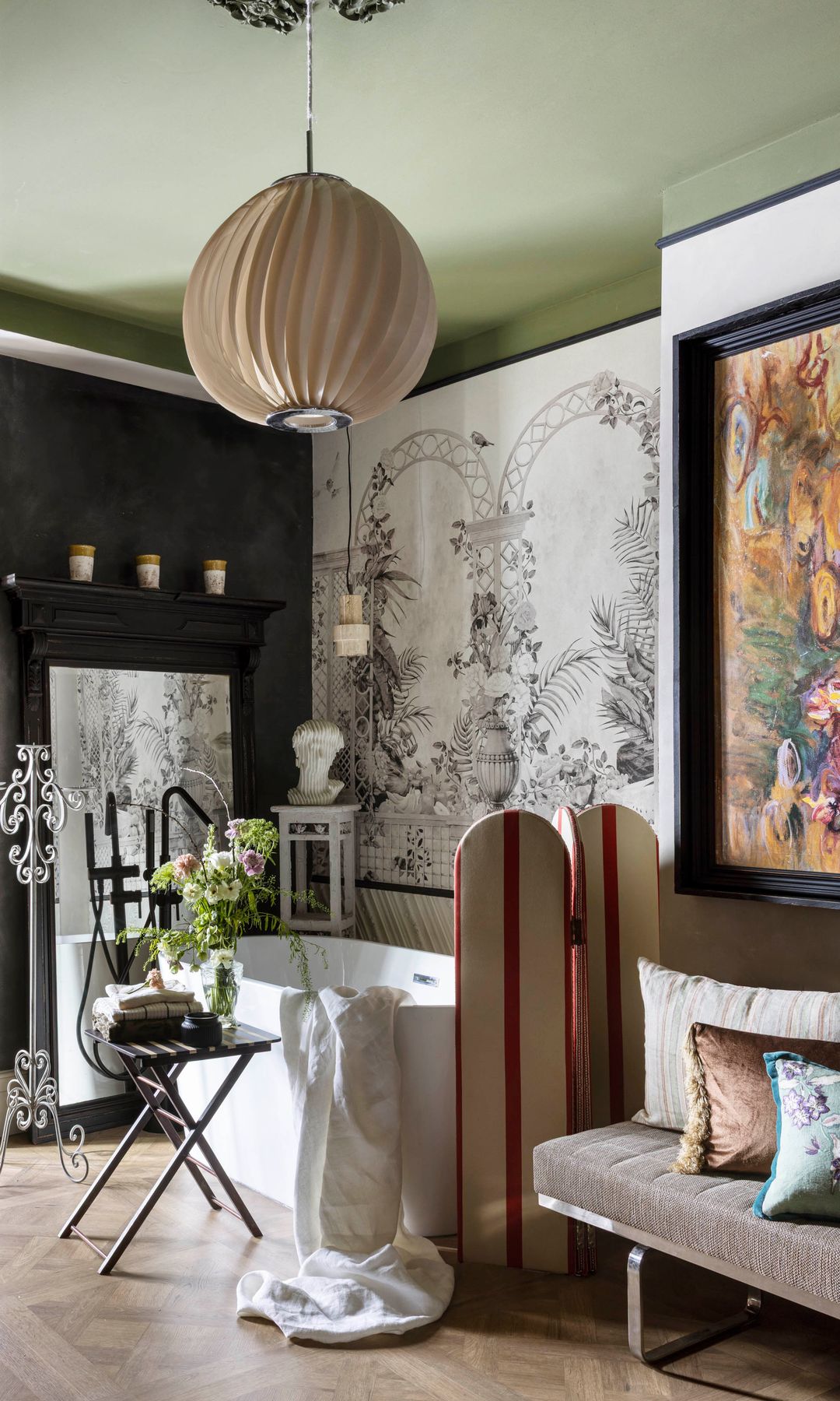 Baño con bañera exenta, mural en la pared, silla con jarrón con flores y libros, banco tapizado, techo de color verde, lámpara de techo con rosetón, biombo