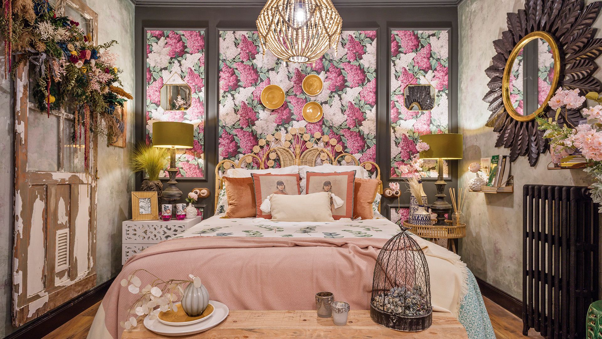 Dormitorio con papel pintado de flores rosas, espejo redondo con marco de hojas, banco a los pies de la cama, lámparas de sobremesa con pantalla amarilla, platos amarillos en la pared del cabecero, lámpara de techo