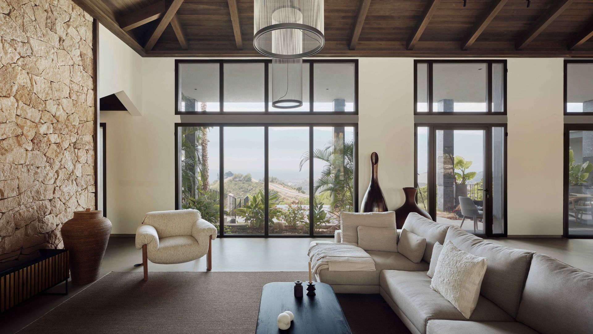 Salón con sofá beige, grandes ventanales que comunican con el jardín, techo de madera, alfombra, butaca blanca, mesa de centro negra, lámpara de techo