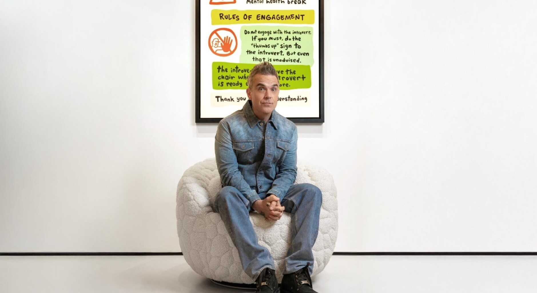El músico Robbie Williams sentado en la butaca blanca junto al manifiesto para introvertidos