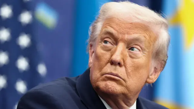 US President Donald Trump, wearing a suit and pictured in front of a US flag, looks to his right.