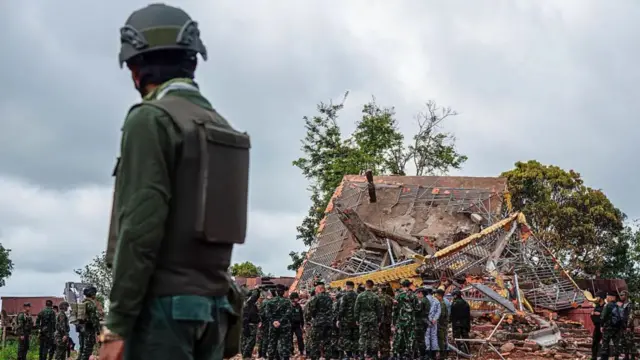 Un soldado camboyano monta guardia cerca de un edificio destruido en la frontera entre Tailandia y Camboya. Observa a un equipo de soldados y funcionarios que inspeccionan el lugar. 