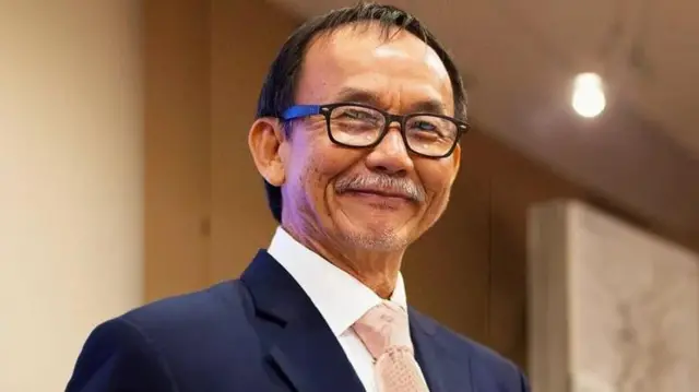El pastor Raymond Koh sonríe a la cámara mientras viste un traje azul oscuro, una corbata rosa y gafas de montura negra.