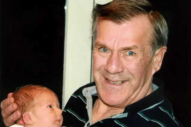 Foto de Ronnie Lockwood (a la derecha) sonriendo mientras sostiene a un bebé. Tiene los ojos azul brillante y mira sonriente a la cámara. Con su mano izquierda sostiene con cuidado la cabeza del bebé.