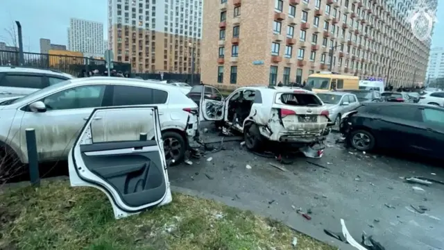 Un auto blanco dañado por la explosión de la bomba. Está rodeado de otros autos en un estacionamiento. 