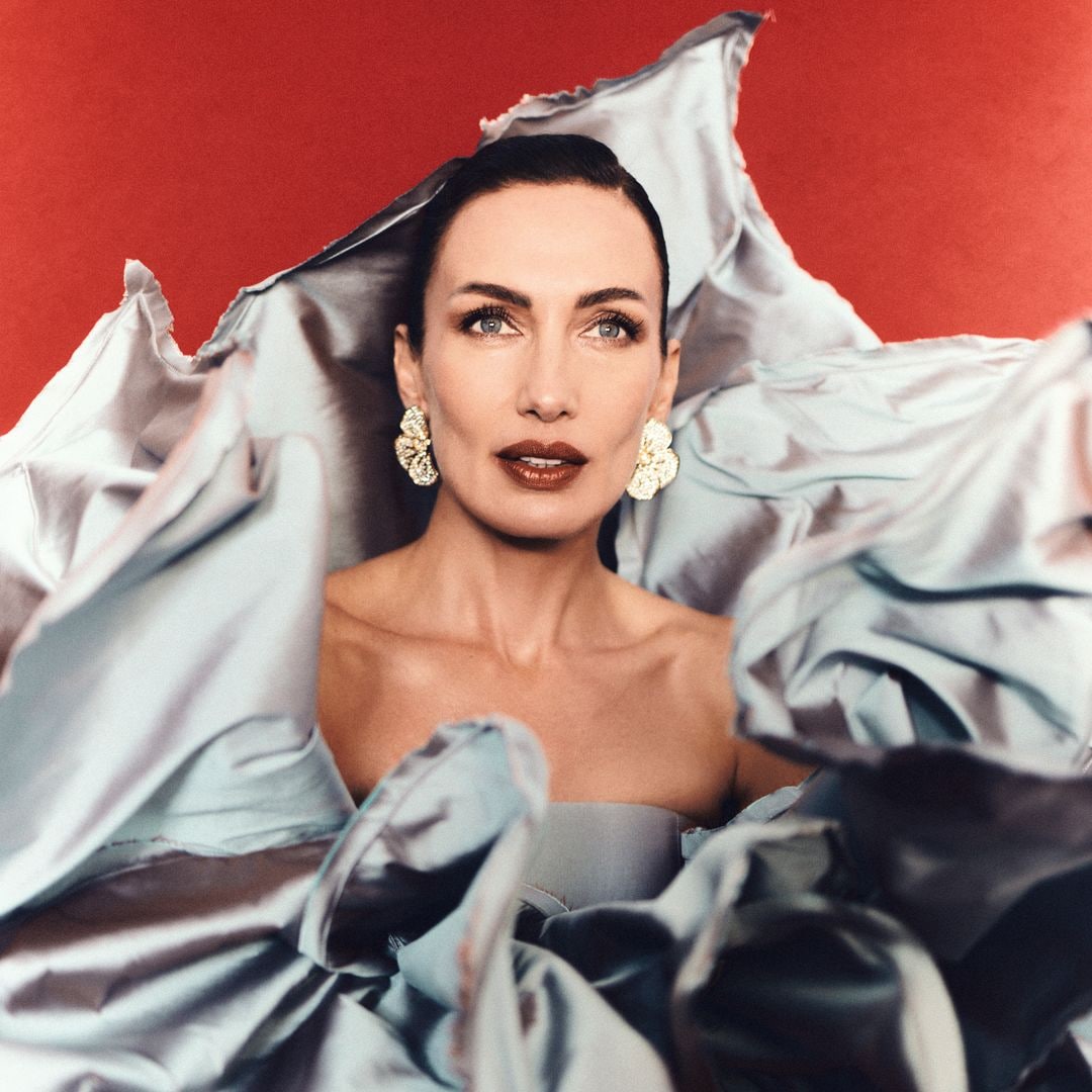 Nieves Álvarez con cabello recogido, aretes grandes, y vestido plateado, posando con fondo rojo