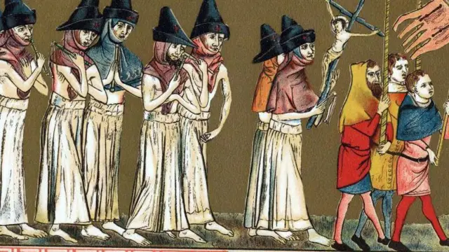 Ocho personas vestidas con túnicas blancas y sombreros puntiagudos caminan descalzas en una procesión detrás de un grupo de tres hombres con ropa colorida. Los hombres llevan banderas.