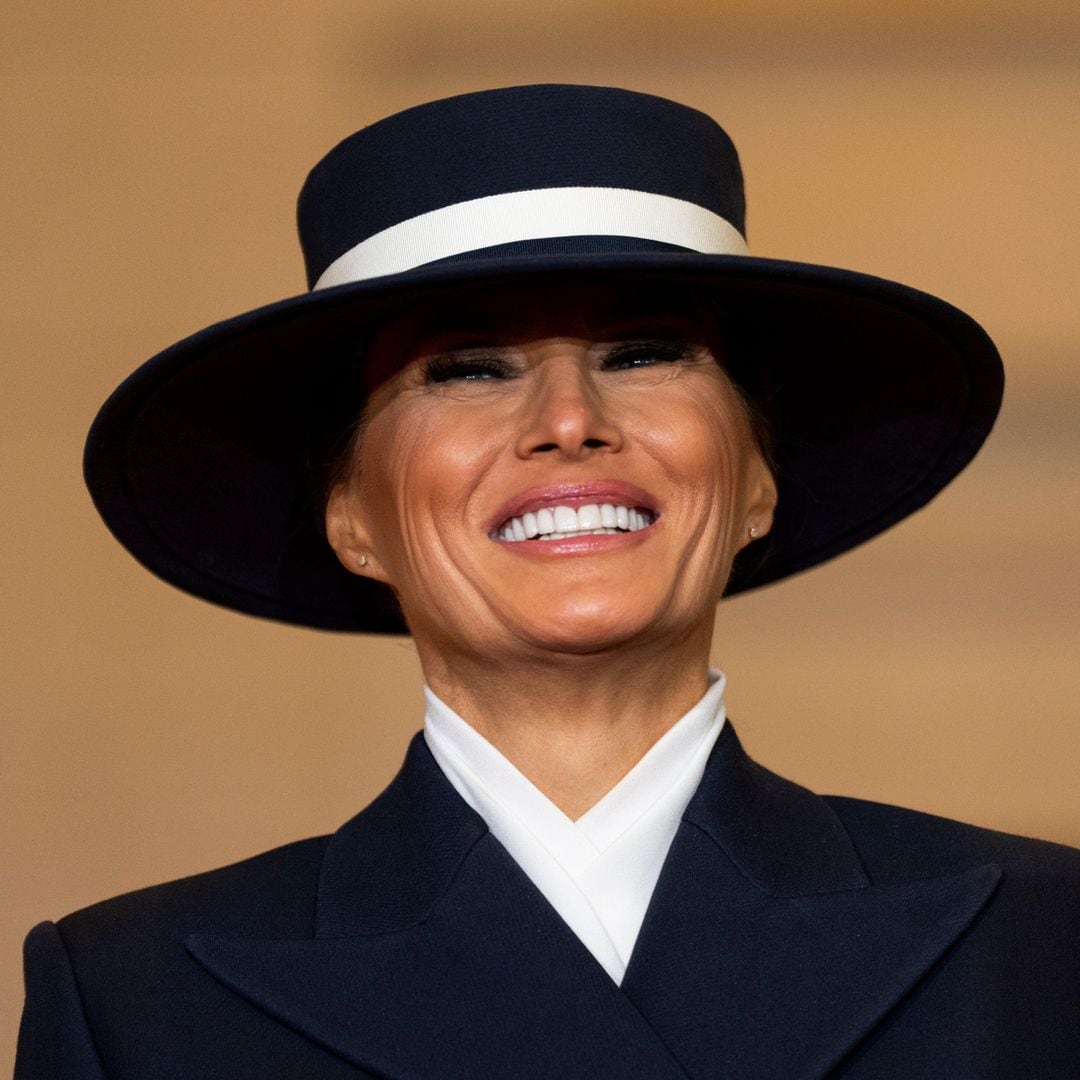 Melania Trump sonriendo, vistiendo traje azul y sombrero negro con banda blanca.