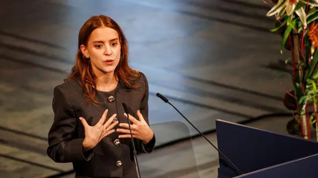 Ana Corina Sosa Machado, hija de María Corina Machado, ofrece un discurso en nombre de su madre, a quien le fue otorgado el premio nobel de la paz. lleva el cabello sobre los hombros y vestuario gris. 