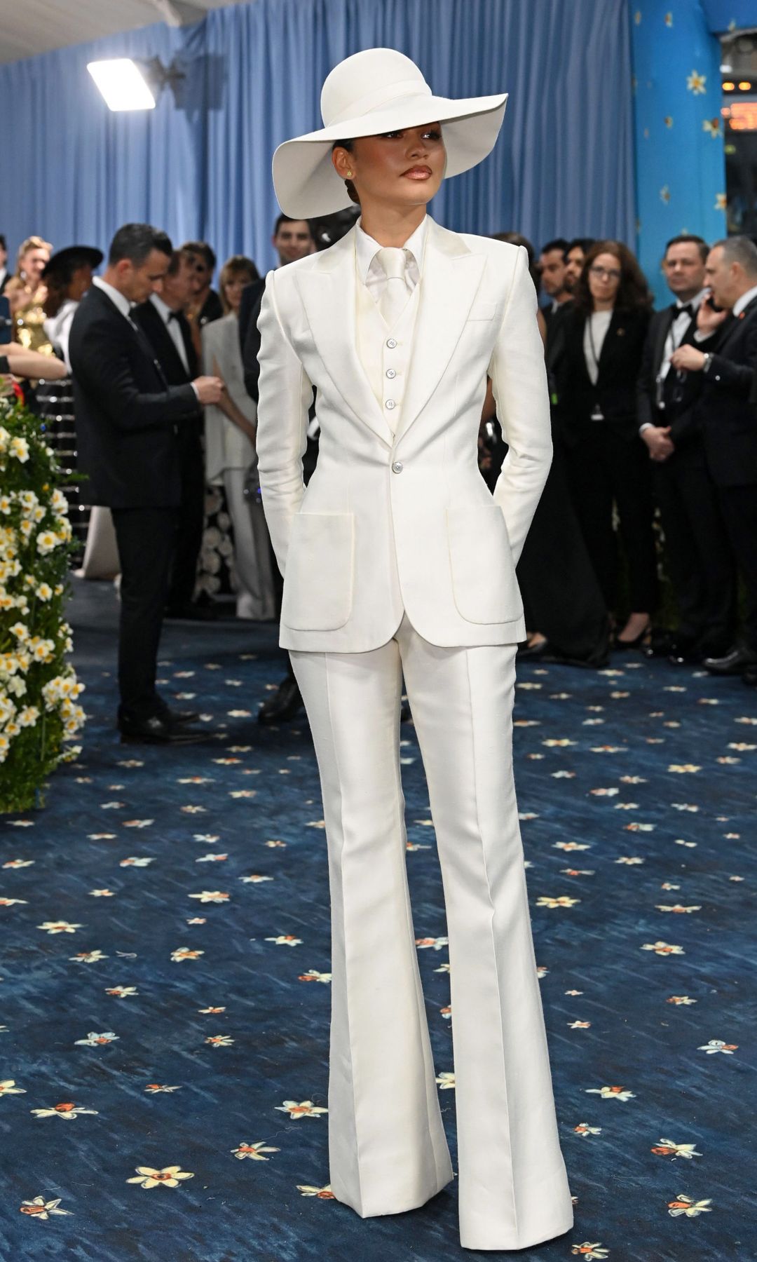 Zendaya en traje blanco, sombrero y corbata, posa en alfombra azul con flores.
