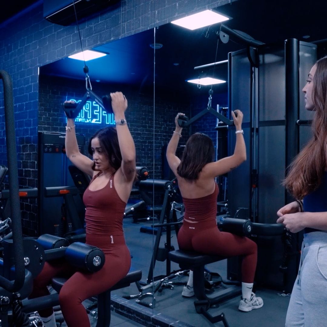 Marina Rivers y su entrenadora en un gimnasio, una sentada con brazos arriba y otra de pie observando.