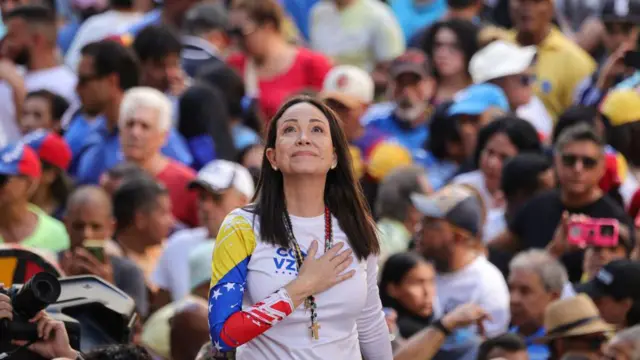 María Corina Machado de pie, con una mano en el pecho, tiene la vista al cielo. Viste una camisa con los colores de la bandera venezolana, azul, amarillo y rojo. También tiene blanco. Detrás de ella se ve una multitud de gente difuminada. 