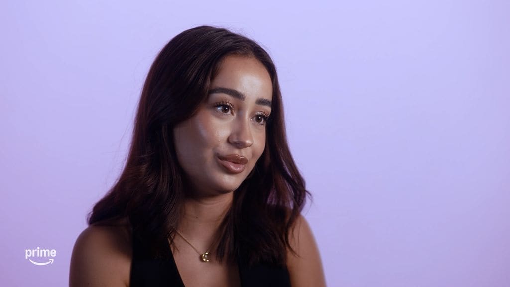 Marina Rivers con cabello largo, usando un collar y una camiseta negra, en un fondo púrpura.