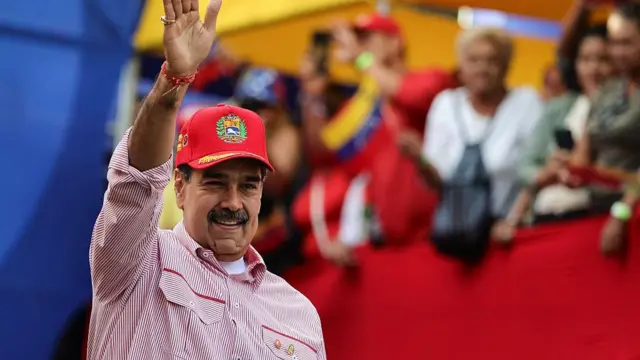 Nicolás maduro saluda con la mano extendida y una sonrisa a un grupo de personas. Tiene una gorra y camisa rojas. 