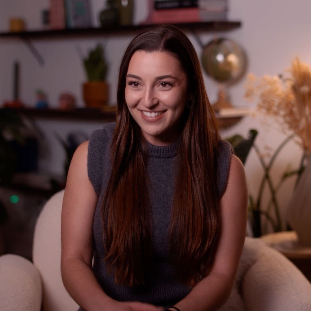 Mujer con cabello largo y sonrisa, sentada en una silla en una sala con decoración acogedora.