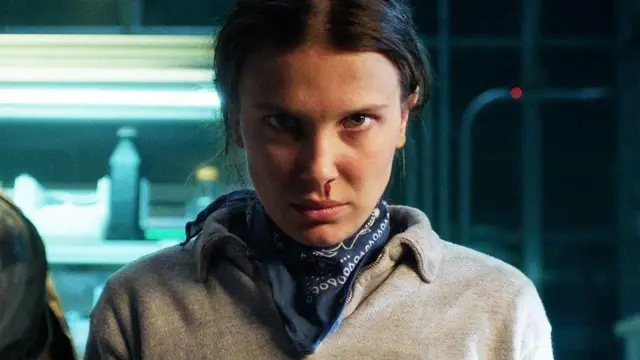 El personaje de Stranger Things, Eleven, una adolescente, mira fijamente a la cámara. Lleva un suéter gris con un pañuelo azul marino estampado alrededor del cuello. Una gota de sangre le cae de la nariz hacia el labio. Tiene la cabeza baja y el cabello oscuro recogido. Detrás de ella, se ven estanterías metálicas iluminadas con luces de neón y llenas de lo que parecen ser botellas.