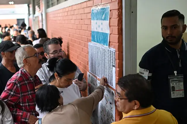 Personas revisando listas de votantes en una pared con bandera de Honduras durante un proceso electoral.