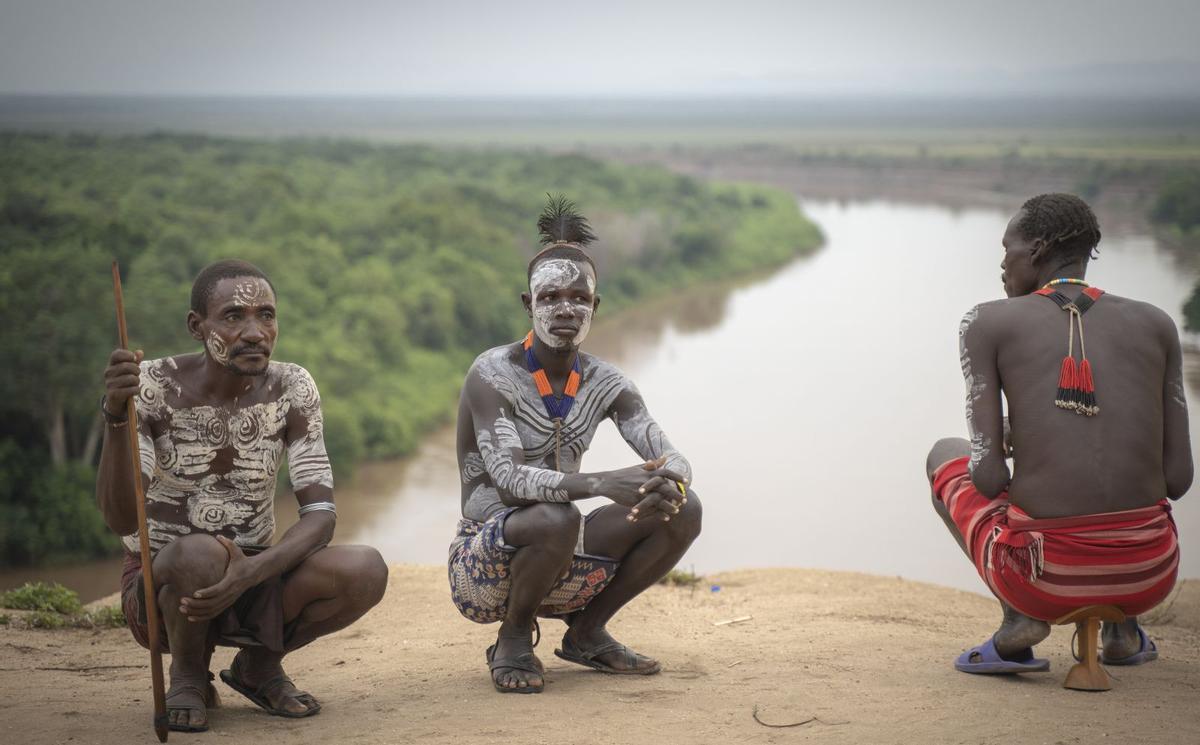 A la izquierda, tres hombres himba, ante el río Omo. En el centro y de arriba abajo, una joven himba, con el cuelo cubierto de collares; dos niños etíopes se protegen del sol con hojas; otro pequeño himba recoge en un cuenco sangre de una vaca para beberla en uno de sus ritos y una mujer etiope, con el pelo rojizo por el óxido de hierro. A la derecha, Emilio Cueto con su amiga inseparable, la cámara de fotos y debajo la mayor duna del mundo, la 45, en Namibia. | emilio cueto/Miki lópez | EMILIO CUETO
