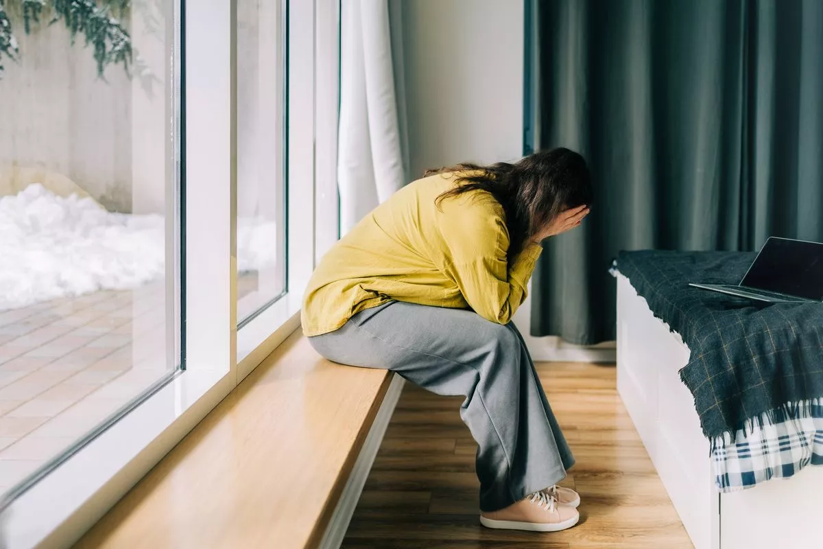  Mujer triste sentada junto a la ventana cubriéndose la cara con las manos