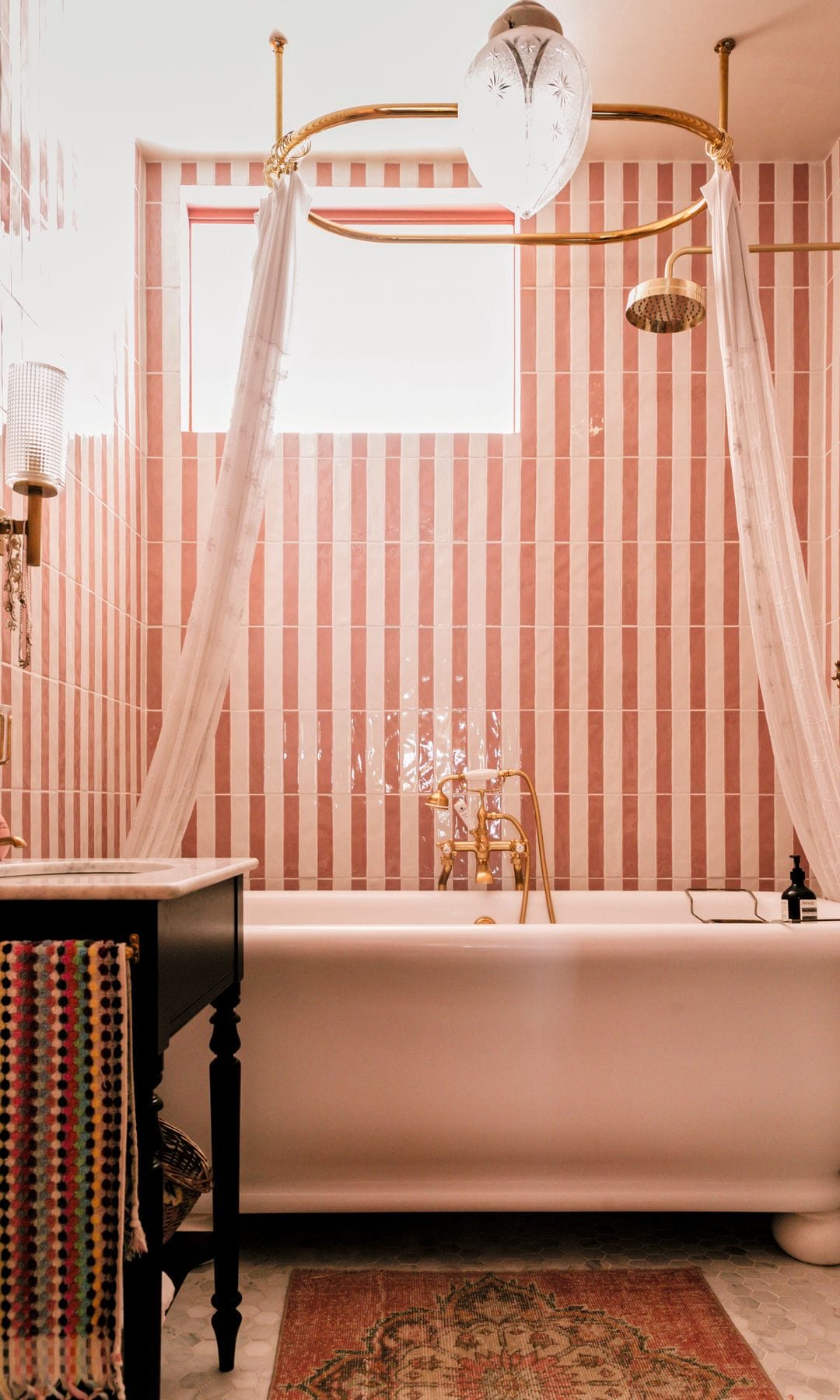 Baño con bañera antigua, cortinas blancas, azulejos rosas y dorados.