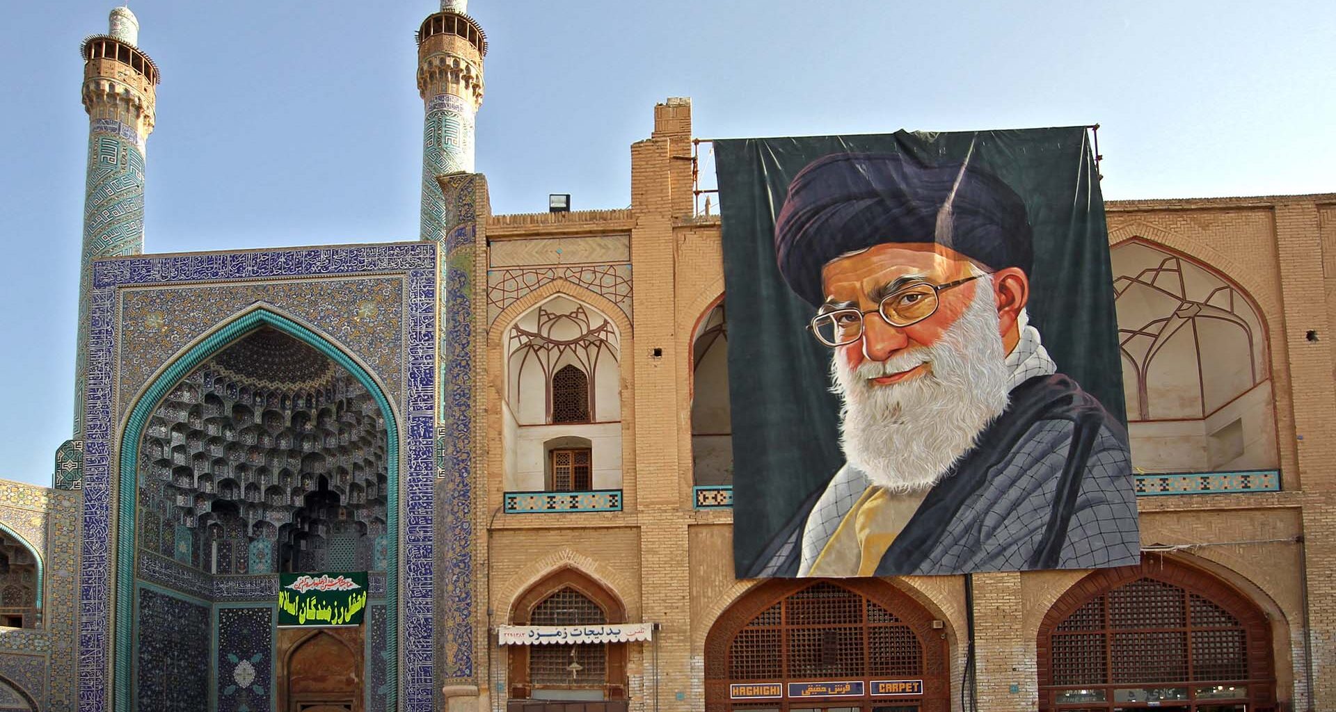 Detalle de la fachada y entrada de la Gran Mezquita de Isfahán (Irán), con su característico arco decorado con azulejos azules y minaretes ornamentados. A la derecha, un gran retrato de Alí Jamenei, líder supremo del país, cuelga sobre un edificio de ladrillo con arcos y motivos geométricos