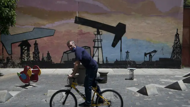 Un hombre en una bicicleta pasa frente a un mural que muestra torres y extracción de petróleo en Caracas en 2020 
