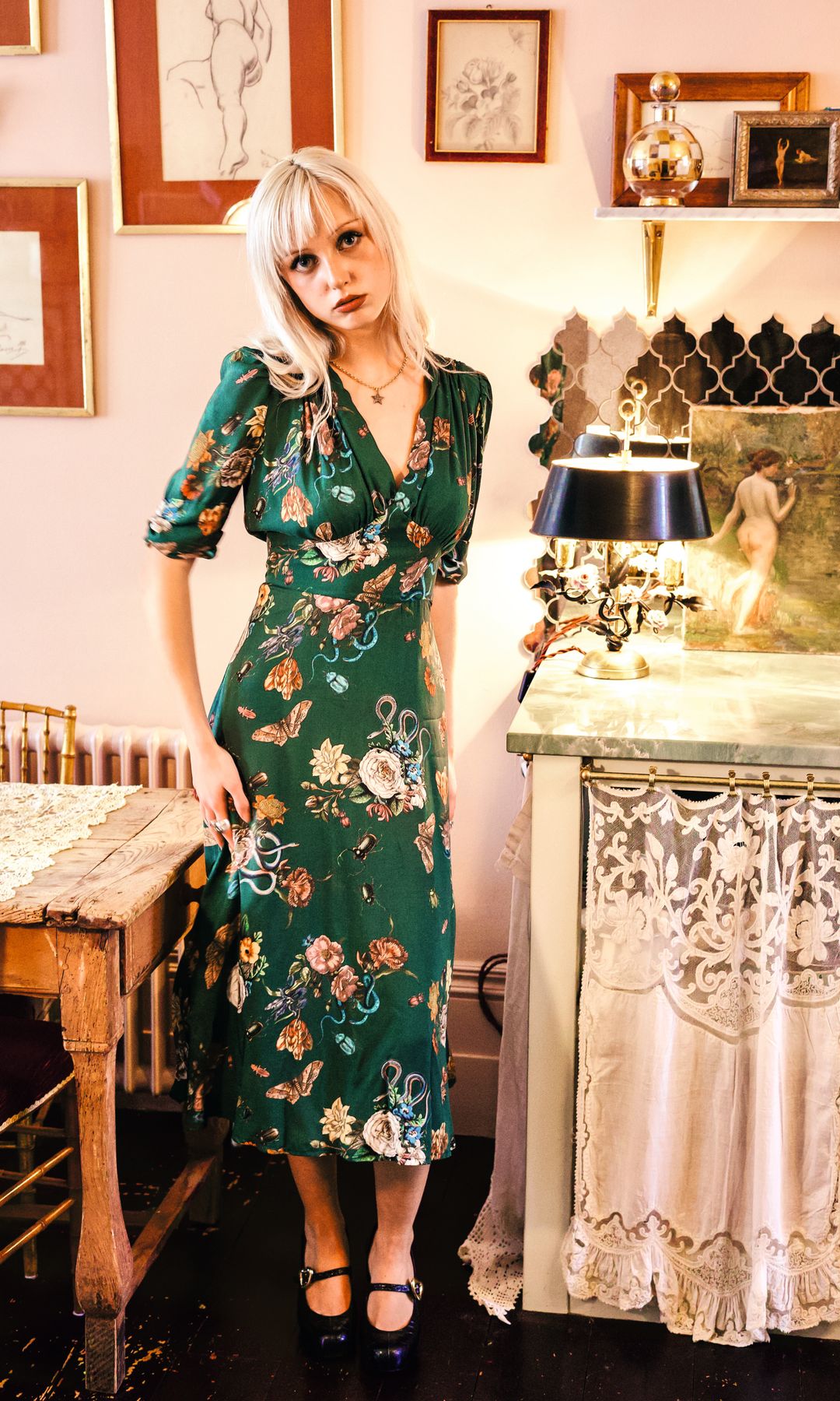 Mujer con cabello rubio en vestido floral verde en una habitación decorada.