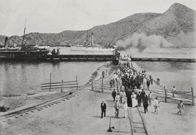 Un grupo de personas caminan por los rieles del ferrocarril y a lo lejos se ven una embarcaciones