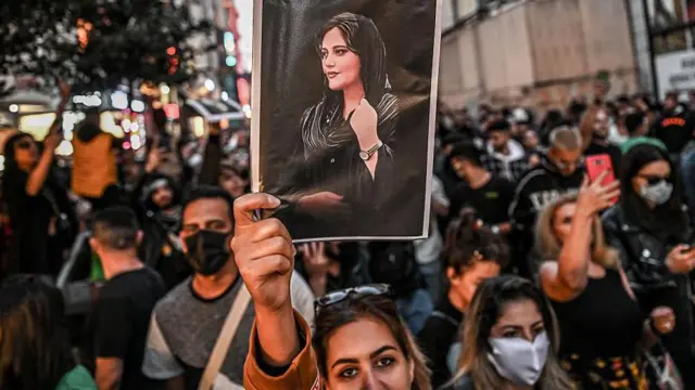Una mujer mira a la camara y alza un retrato de la joven Mahsa Amini, en la que lleva velo. 
