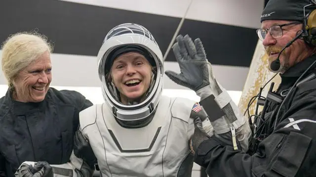 La astronauta de la NASA Zena Cardman, con un traje espacial blanco, saluda sonriente mientras dos personas la ayudan a salir de la nave espacial SpaceX Dragon Endeavour