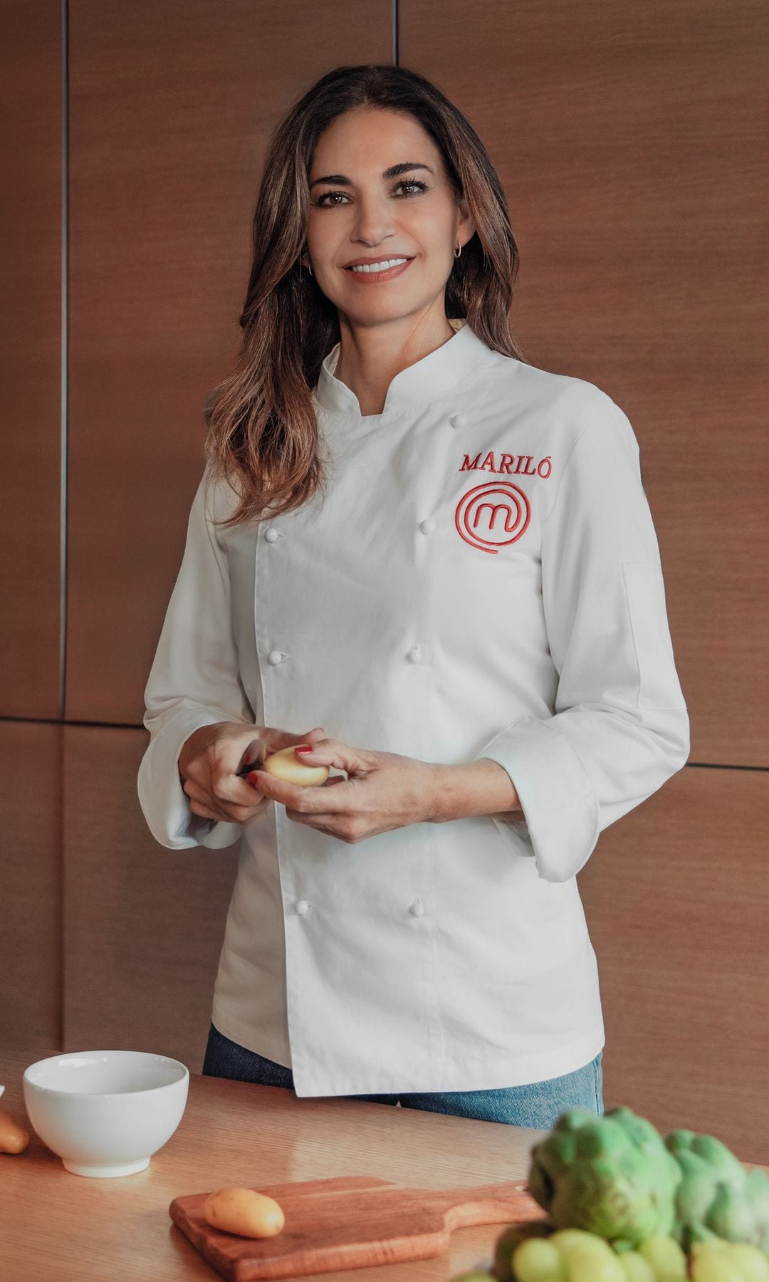 Mariló Montero en uniforme de chef con logo 'M', sosteniendo un alimento, rodeada de ingredientes.