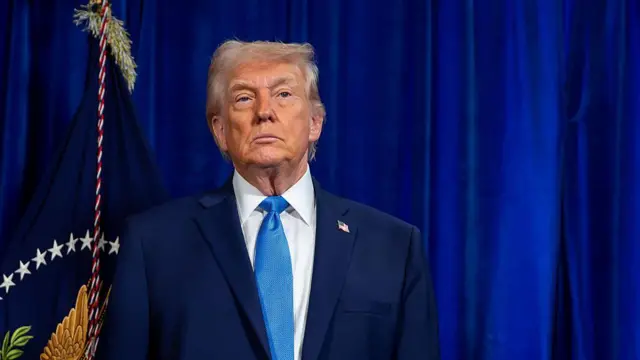 Imagen de donald trump. Viste un traje azul y corbata del mismo color. Está de pie, con rostro serio. 