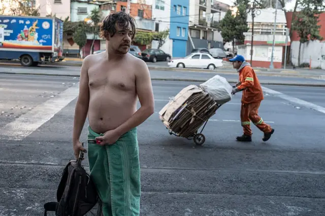 Un hombre en toalla en la mitad de la calle mientras detrás pasa un recolector de cartón