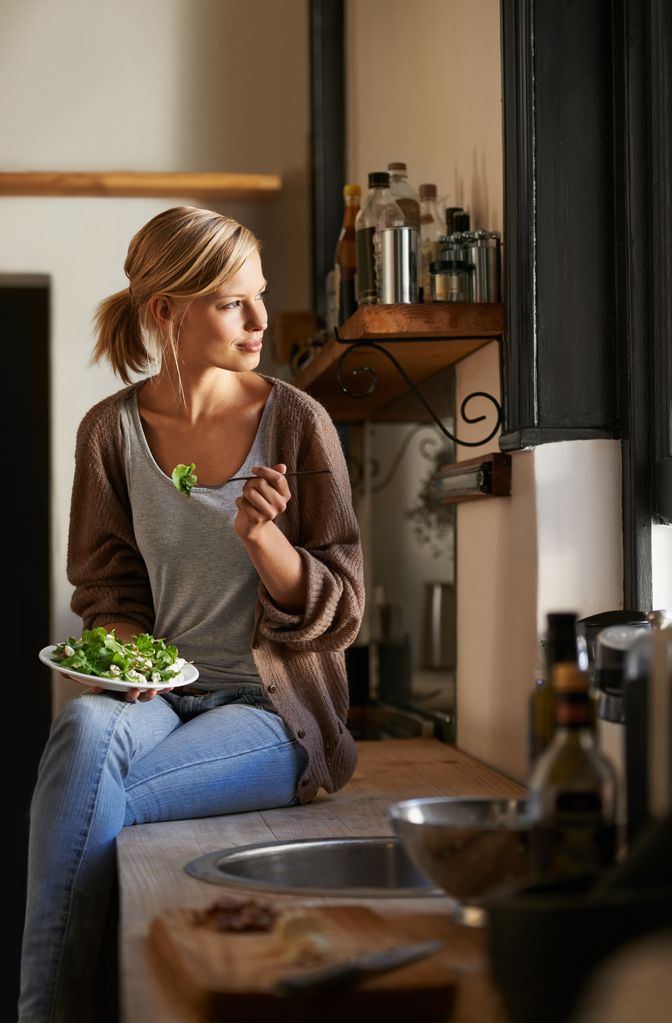 Mujer en la cocina, comiendo ensalada con tenedor, sentada en la encimera.