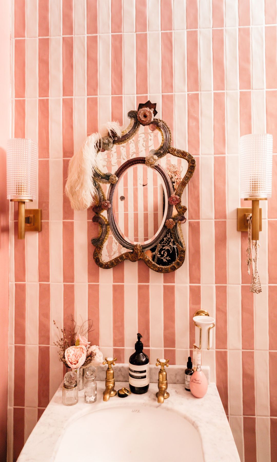 Baño con espejo vintage, lámparas de pared, y accesorios de baño en una encimera de mármol.