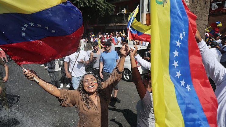 Una mujer con la bandera de Venezuela pintada en la cara sostiene dos banderas y sonríe ampliamente.