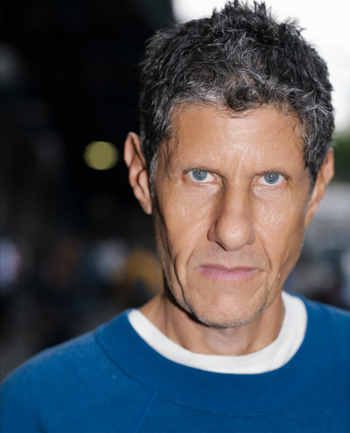 Primer plano de un hombre de cabello gris y ojos azules, con camiseta azul sobre una camiseta blanca de cuello redondo