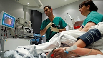 Un médico con guantes manipula un endoscopio durante un procedimiento en una sala de hospital, con un monitor mostrando imágenes internas y una enfermera observando al paciente