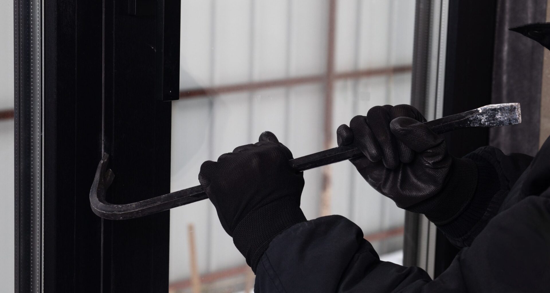 Eine vermummte Person mit schwarzen Handschuhen versucht mit einem Brecheisen eine Tür oder ein Fenster aufzuhebeln. Das Bild symbolisiert physische Einbrüche und Gewaltkriminalität im Zusammenhang mit Krypto-Überfällen.