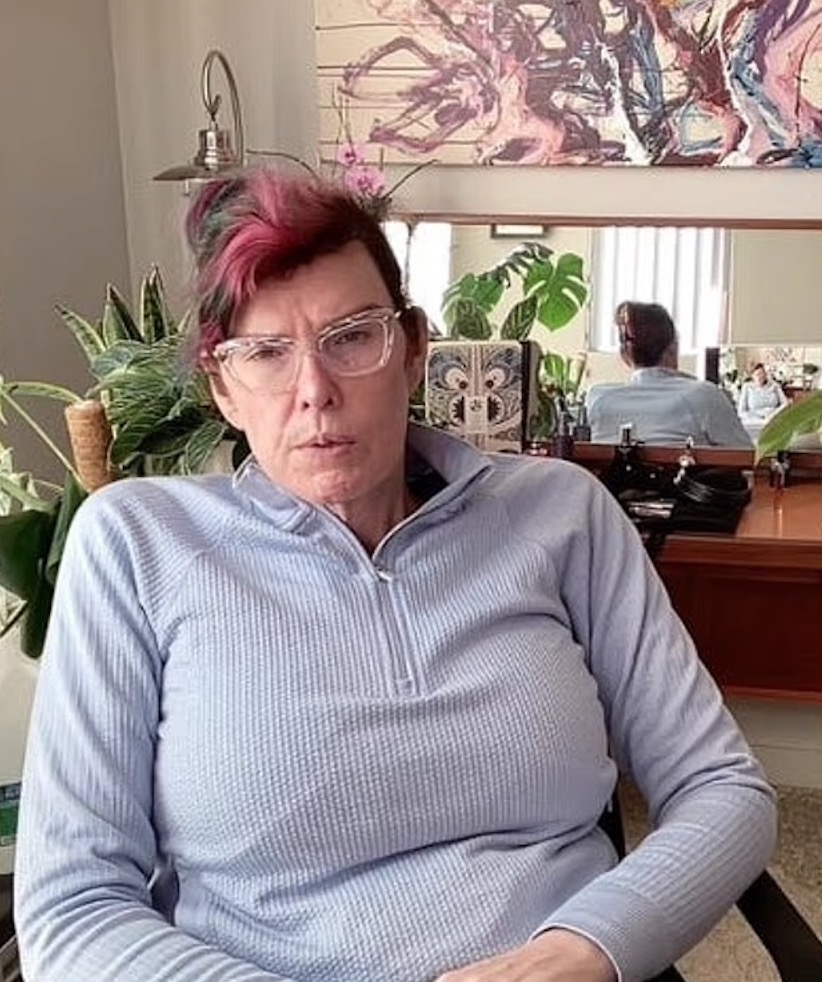 Jane-Michelle Arc, a trans woman with pink hair and glasses, sitting in a light blue top.